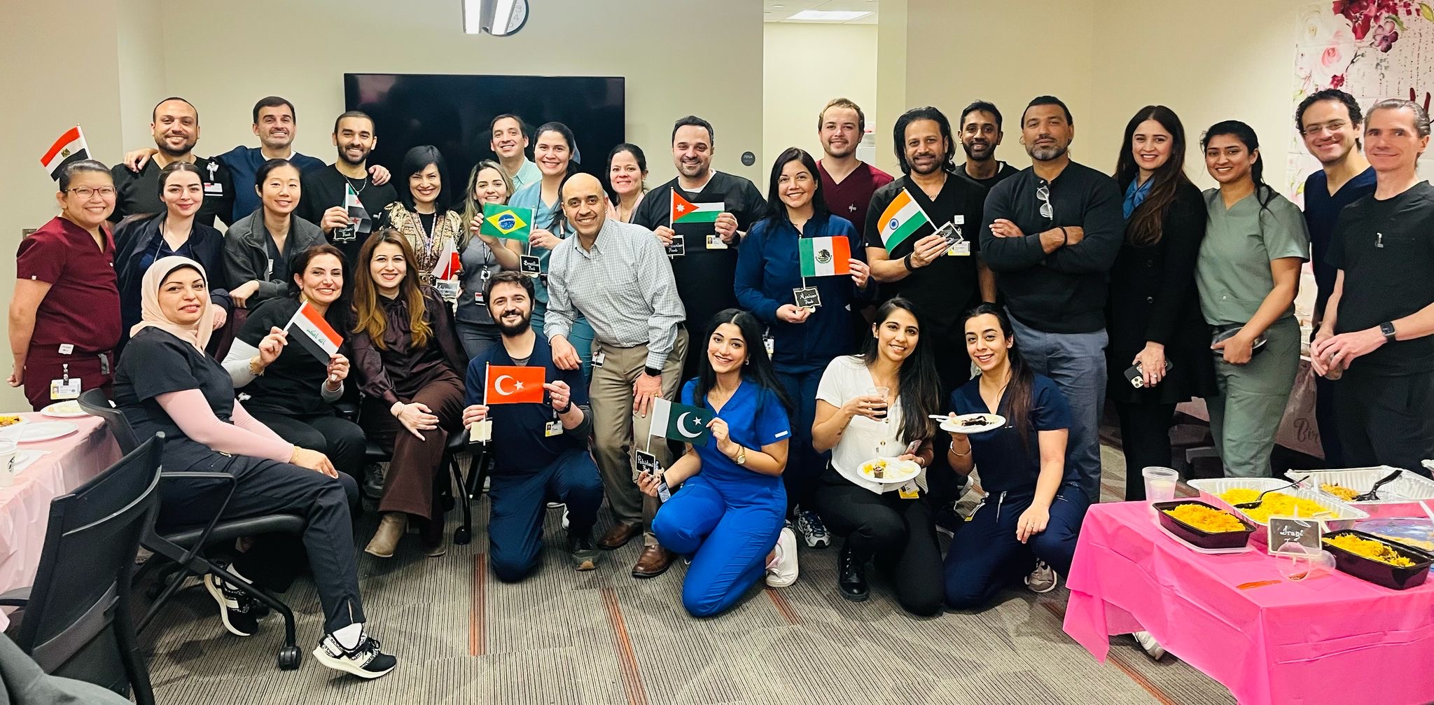 a big group of people in scrubs stands together, with a few holding up flags from various countries. A table of foods from different cultures is seen in the corner. Dr. Baig kneels in the middle holding the Pakistan flag.