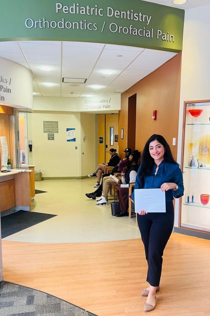 Dr. Baig holds up an award certificate in front of a sign that reads "Pediatric Dentistry, Orthodontics / Orofacial Pain"
