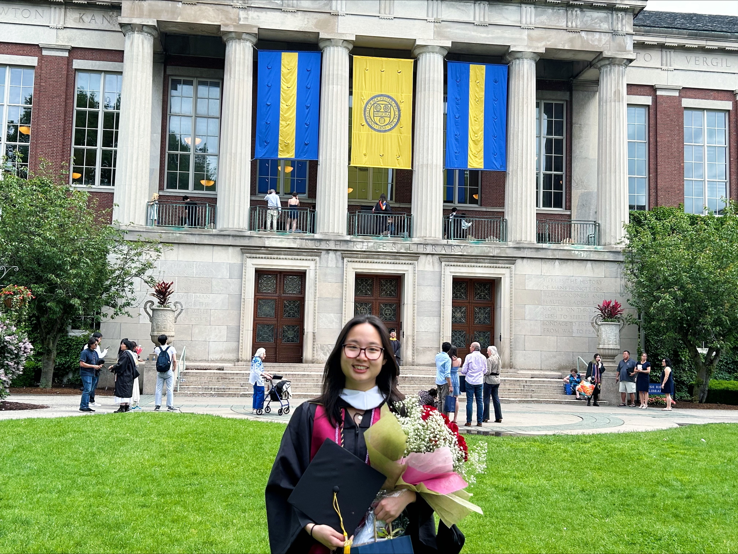 Claire Lee stands in front of Rush Rhees library at the University of Rochester, wearing a graduation robe and holding flowers.