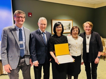 5 people in formal attire stand in a line. The woman in the middle holds up a diploma.