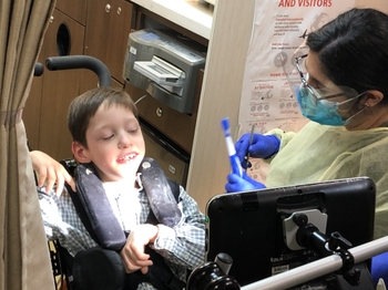 a dentist in gloves, mask, and a yellow plastic gown stands next to a child in a wheelchair and holds a toothbrush and dental mirror.