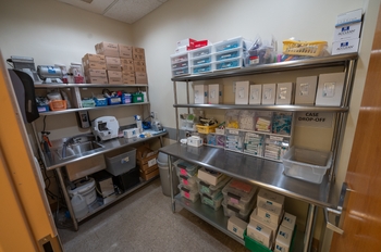 a small room with 2 shiny metal benches with shelving on top with lots of boxes of storage. One bench contains a sink, and has multiple new small machines. Old machines can be seen on a shelf above.