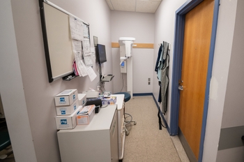 an x-ray machine is seen in the back of a small closed off area, facing a side wall. There are 2 cabinets and a cork board to the side of the x-ray machine. A door to the right has blue trim.