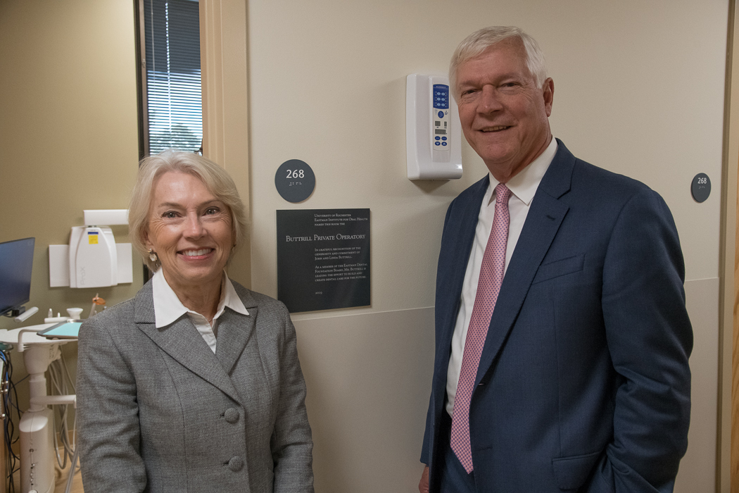 A man and a woman in suits stand on either side of a plaque reading "Buttrill Private Operatory". Behind them is a door into a small room in which a dental chair can be seen.