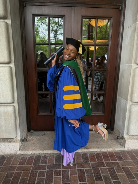 Student stands in academic regalia, smiling with diploma