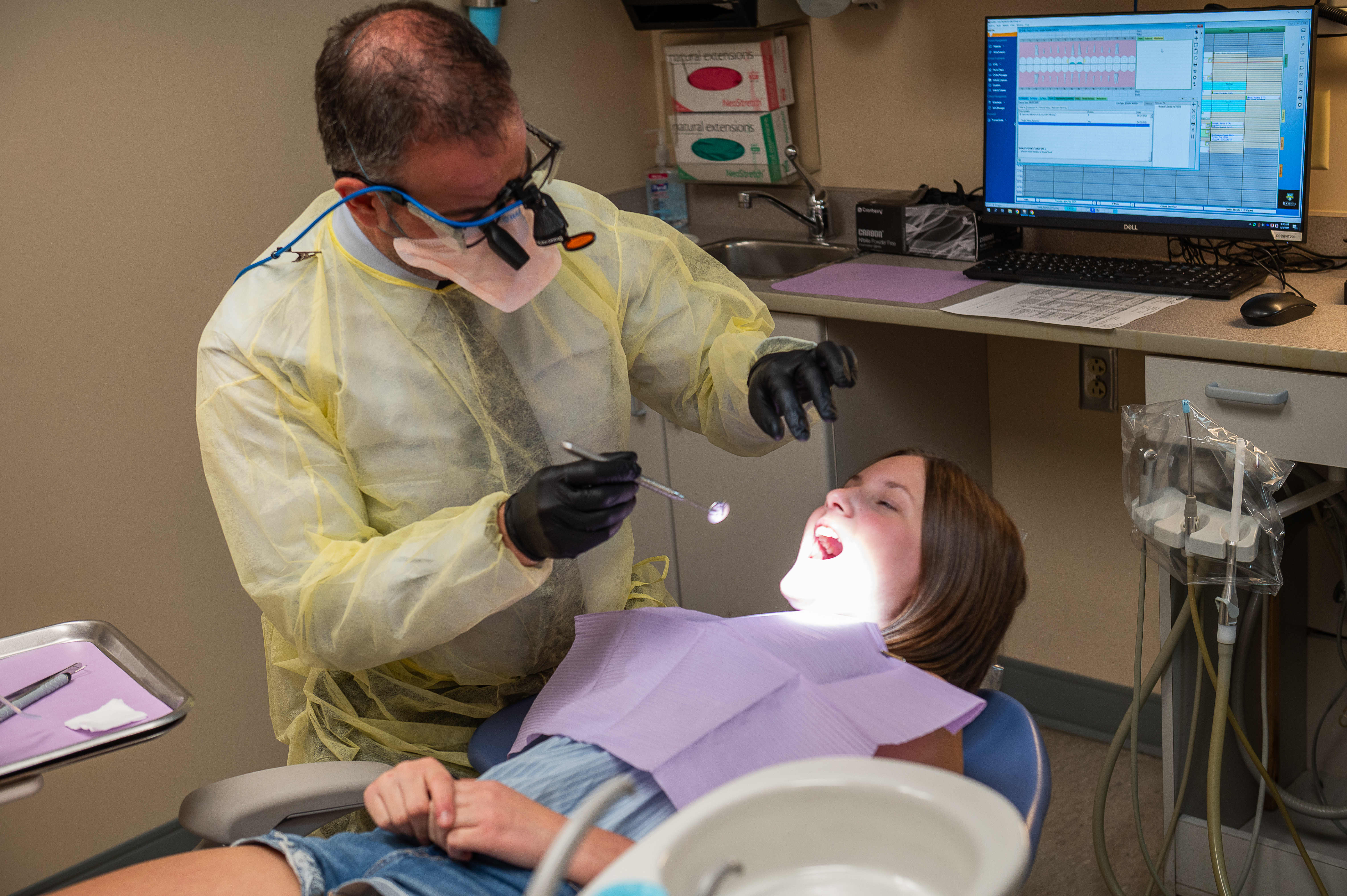 A girl is leaned back in a dental chair while a dentist uses a light and mirror to check her teeth.