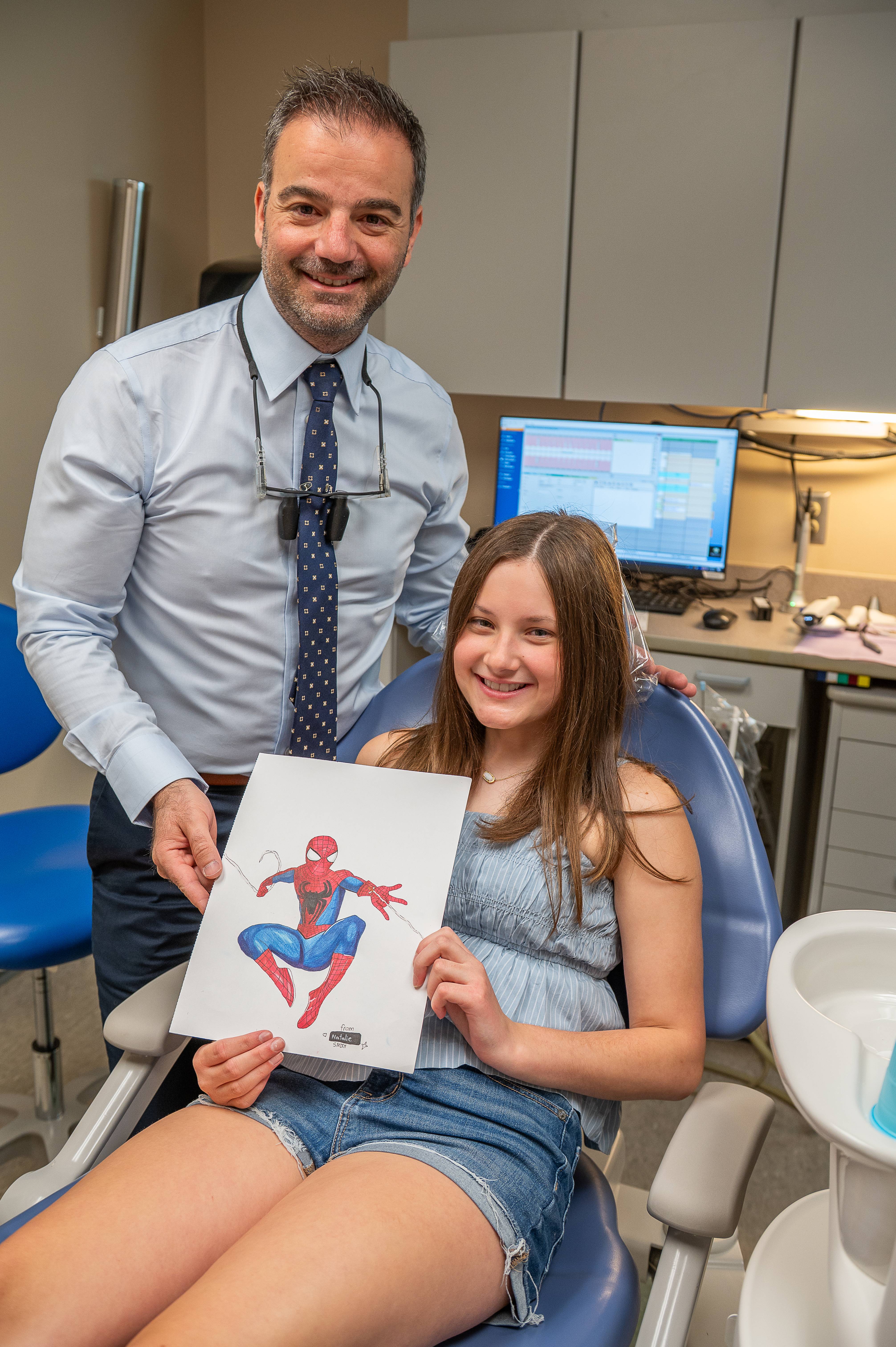 a girl sits in a dental chair and proudly holds up a drawing of Spiderman. A dentist stands beside her smiling.
