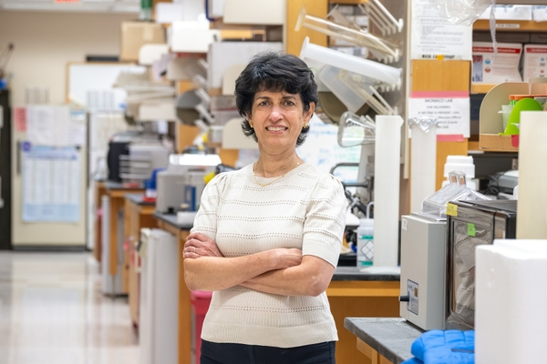 Tanya Mayadas stands with her arms crossed and a research laboratory can be seen behind her.