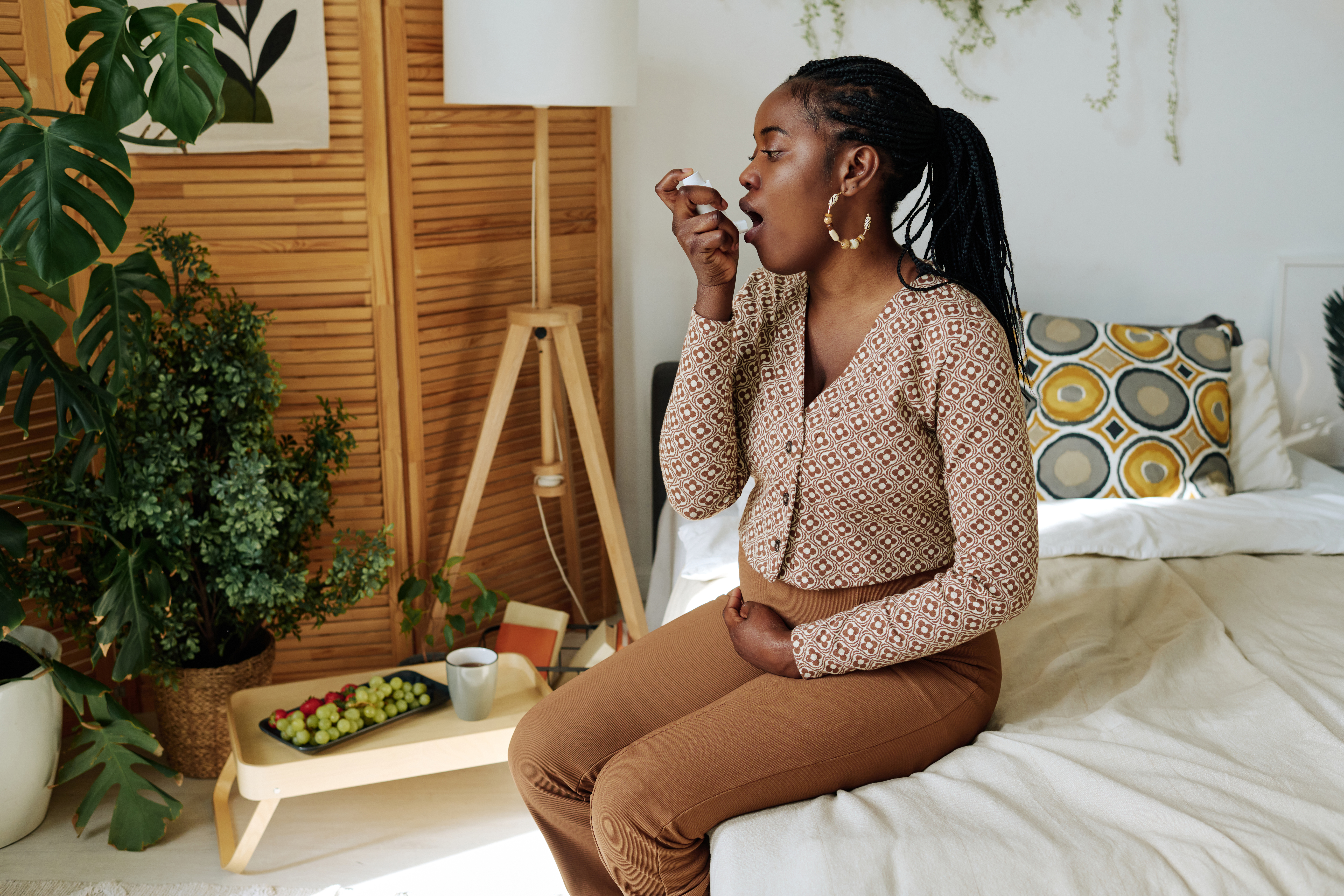 A pregnant person uses an inhaler while sitting on a bed