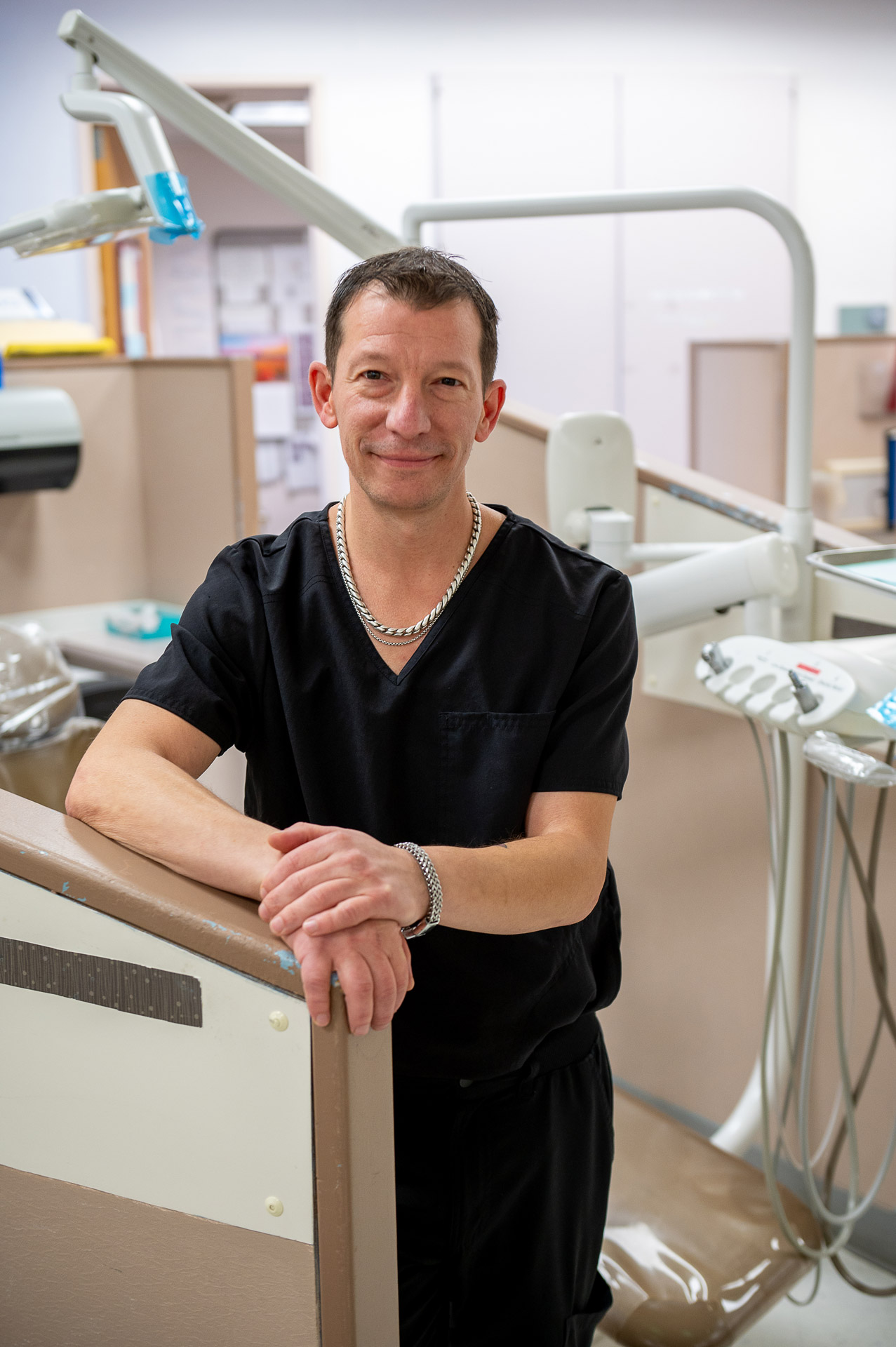 Brian DeWind, standing in the dental clinic wearing black scrubs.