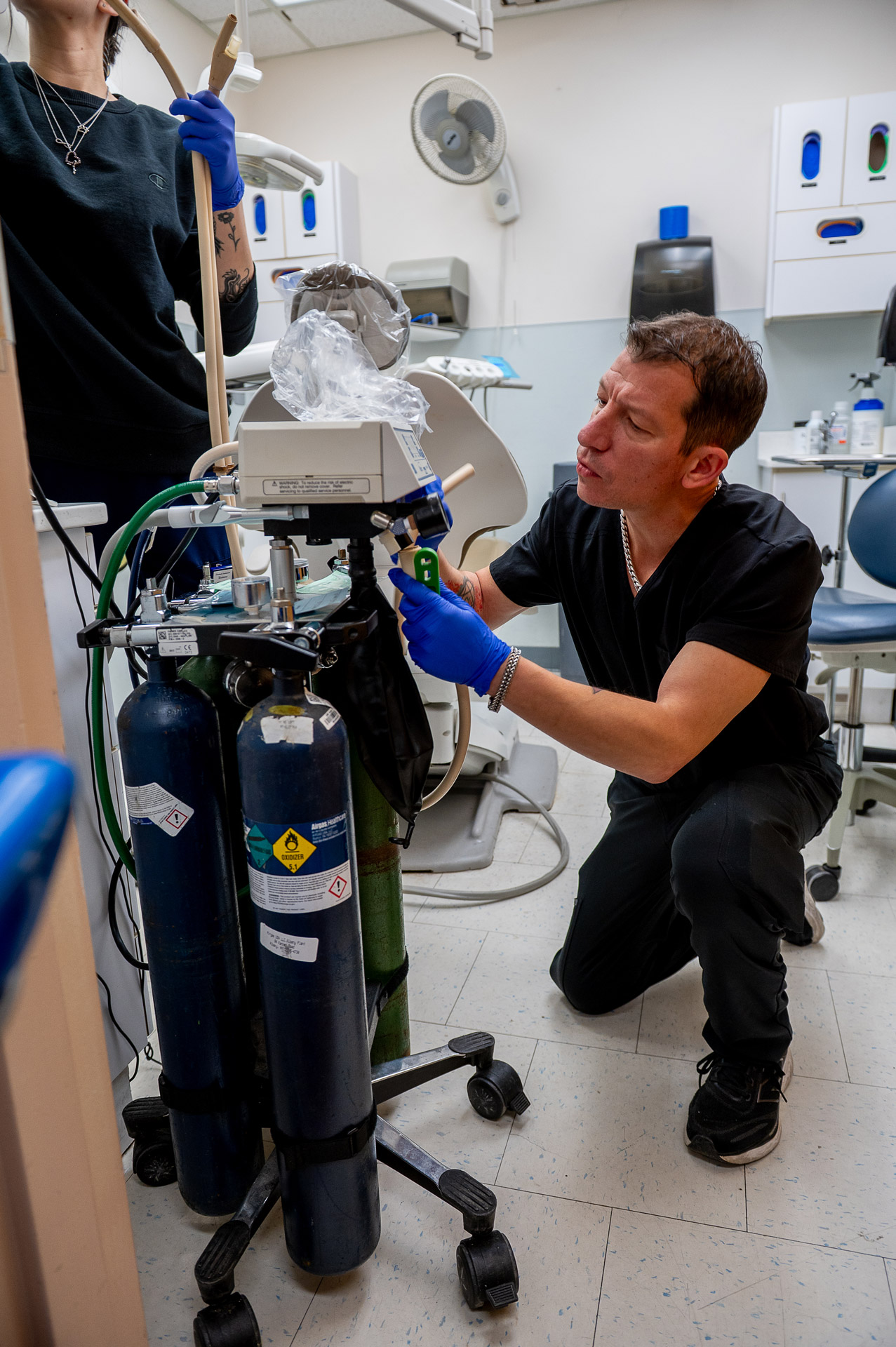 Brian kneels in the dental clinic, working on a device that is hooked up to canisters of gas.