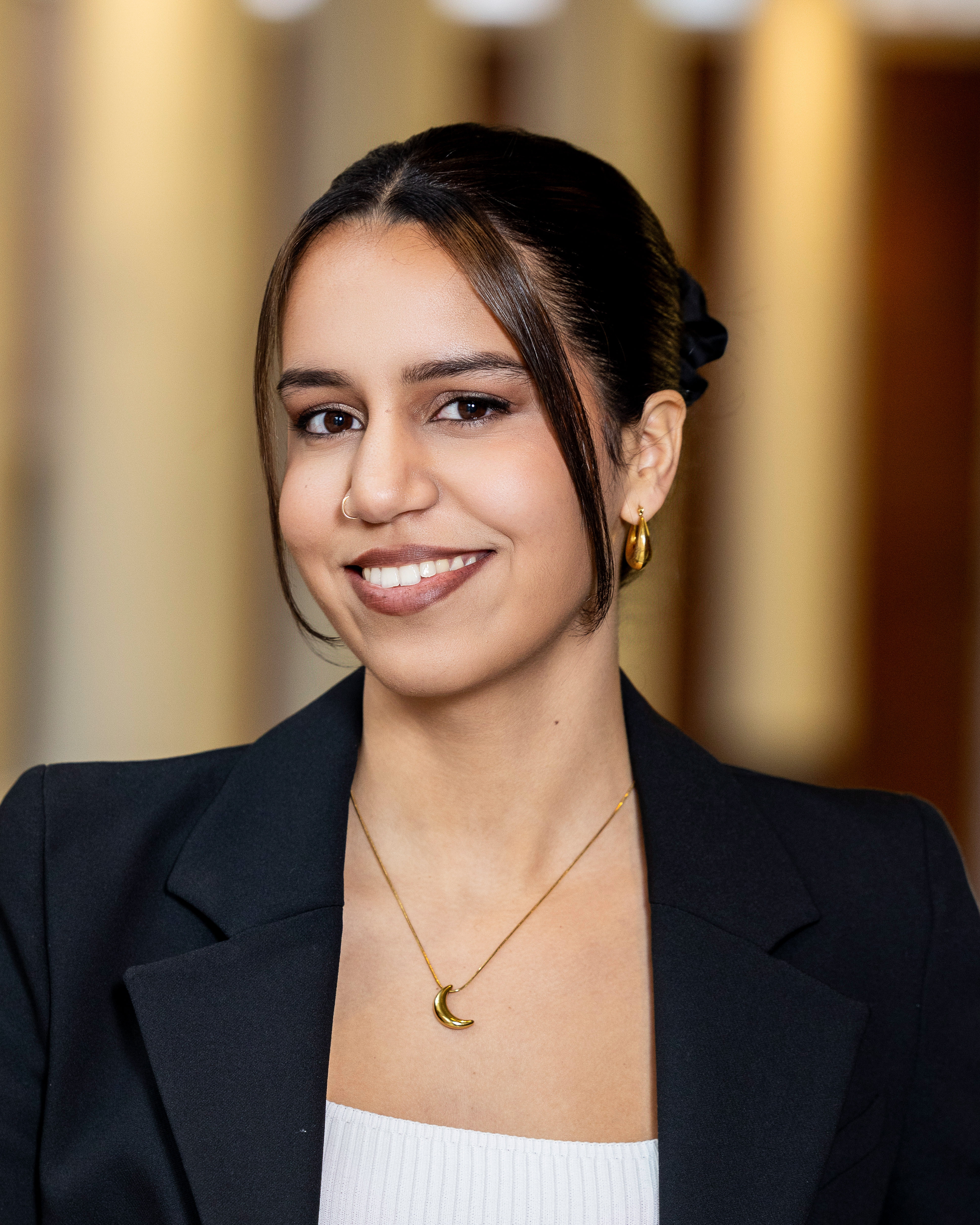 Women with dark hair neatly pulled back, she is smiling and wearing a black blazer and gold jewelry
