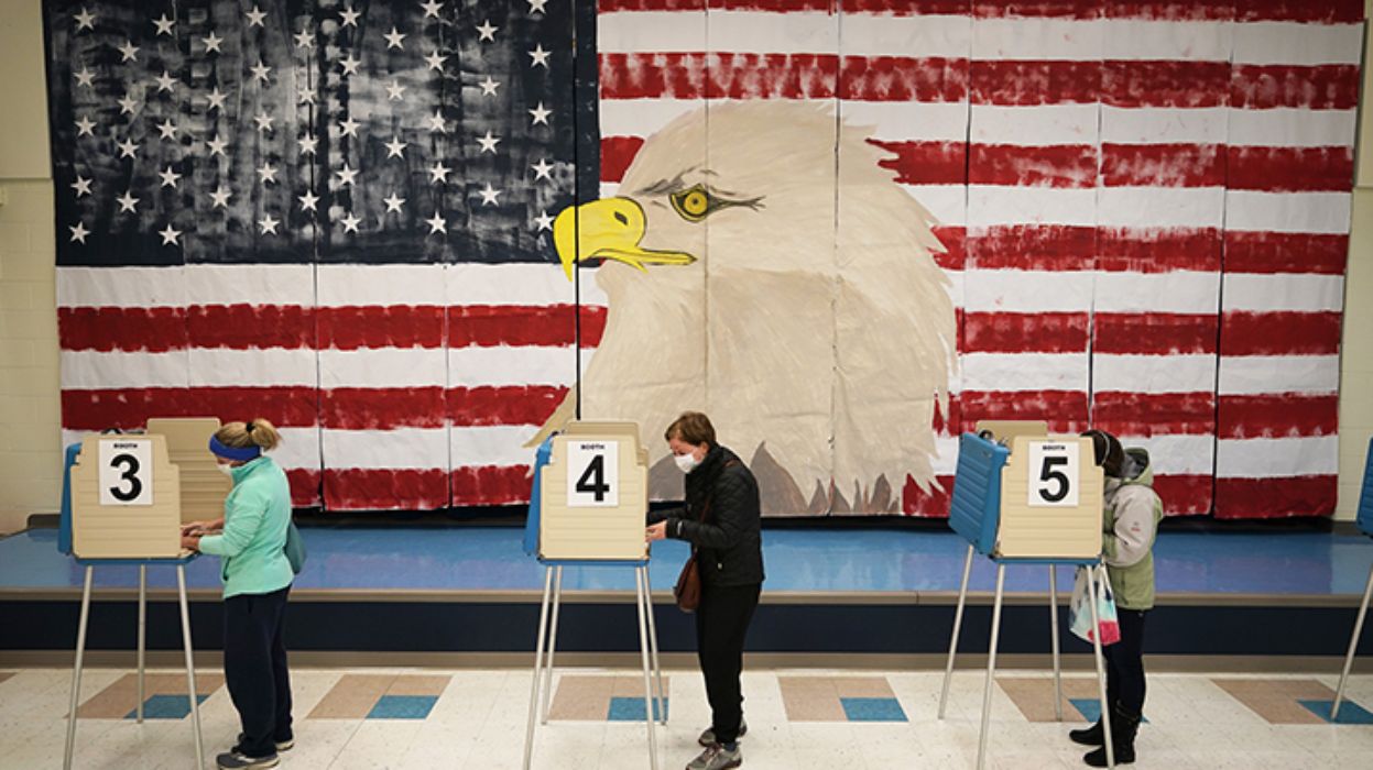 three people voting in a gymnasium with a giant painting of an American flag with an eagle head on the wall to their side