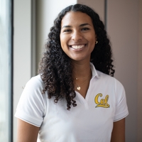 Sydney Roberts, the new ASUC president, poses for a casual portrait. She has a wide smile, a white polo shirt with Cal written on it, gold jewelry and long curly hair.