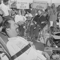 black-and-white photo of people in wheelchairs at a rally holding signs that read "I'm crippled ... not dead" and "Handicapped Americans: A natural resource."