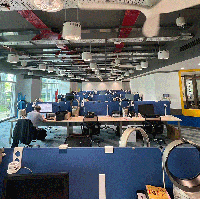 A photo of a brightly-lit open office space. Each desk is equipped with a tower fan.