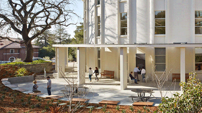 An image of the outside of the Simons Institute for the Theory of Computing at UC Berkeley on a sunny day. Groups of people are pictured talking, studying and walking by the building.