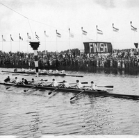 A black and white image of boats being rowed in a race as they cross under a sign that says "finish"