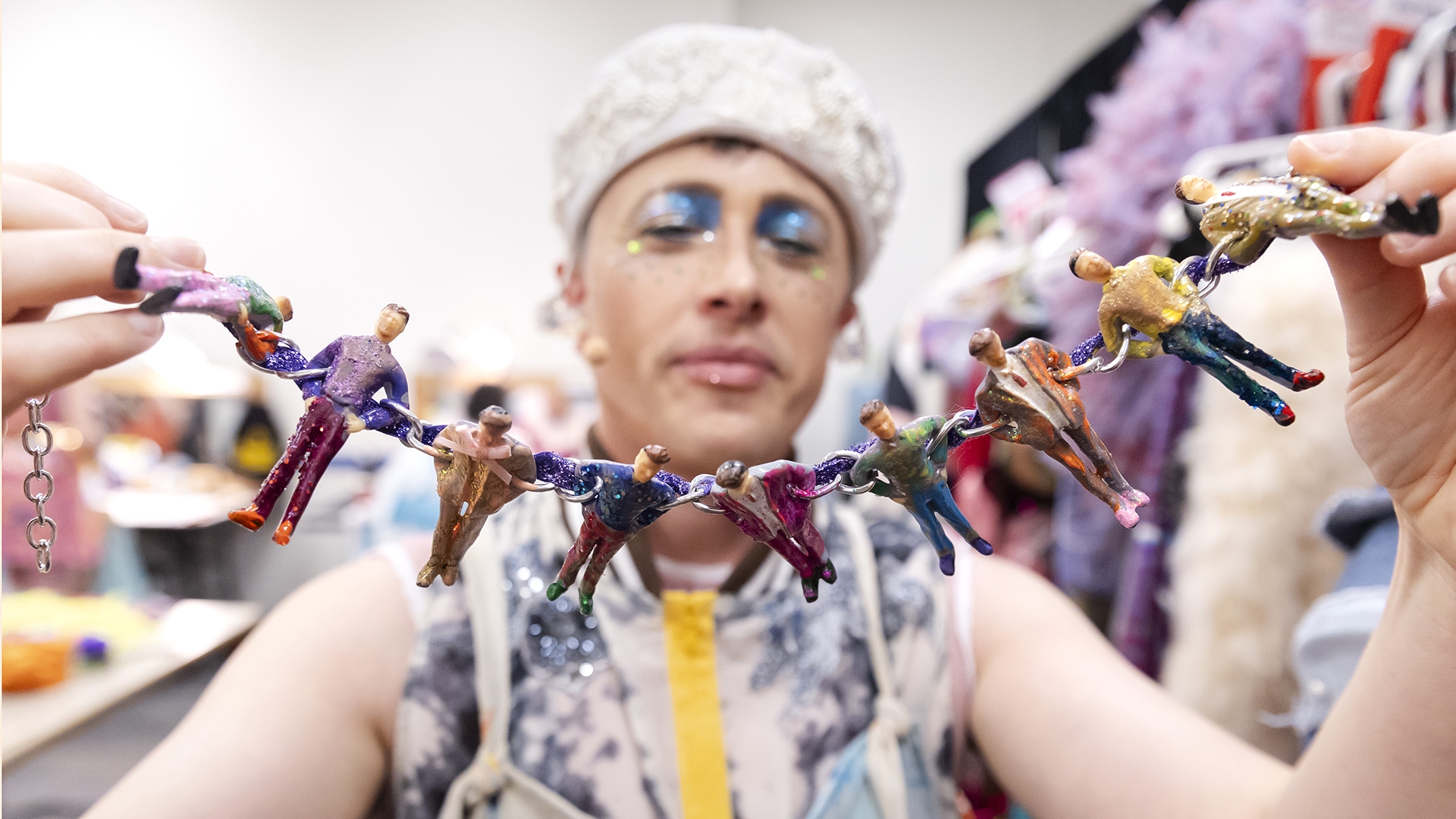 a person holds up a necklace made up of miniature men wearing a variety of colorful, but plain outfits that he will wear in his performances