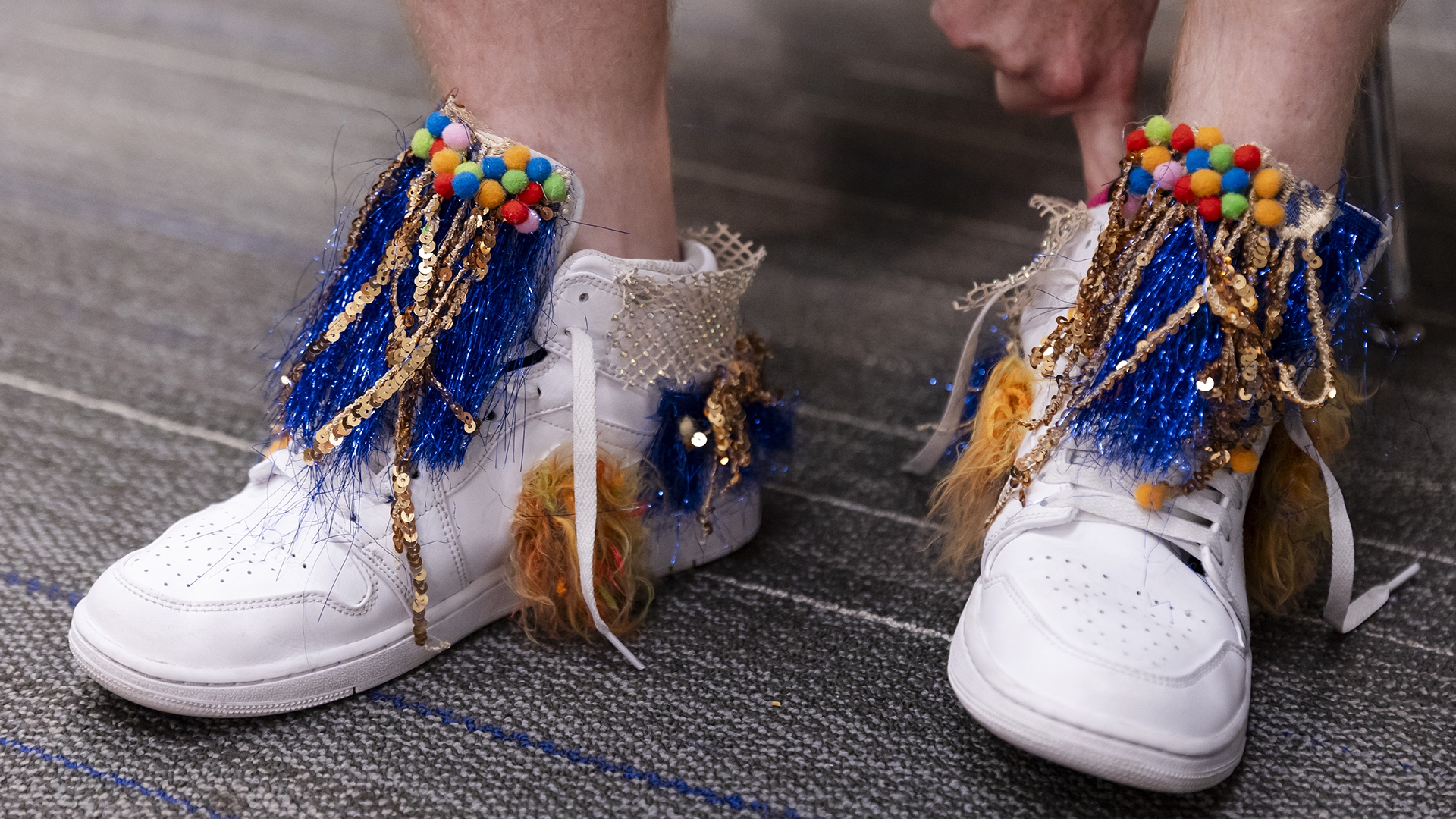 A closeup of a person's feet wearing white hightop shoes with blue and gold tassels hanging from both tongues and little multicolored fabric balls at the top.