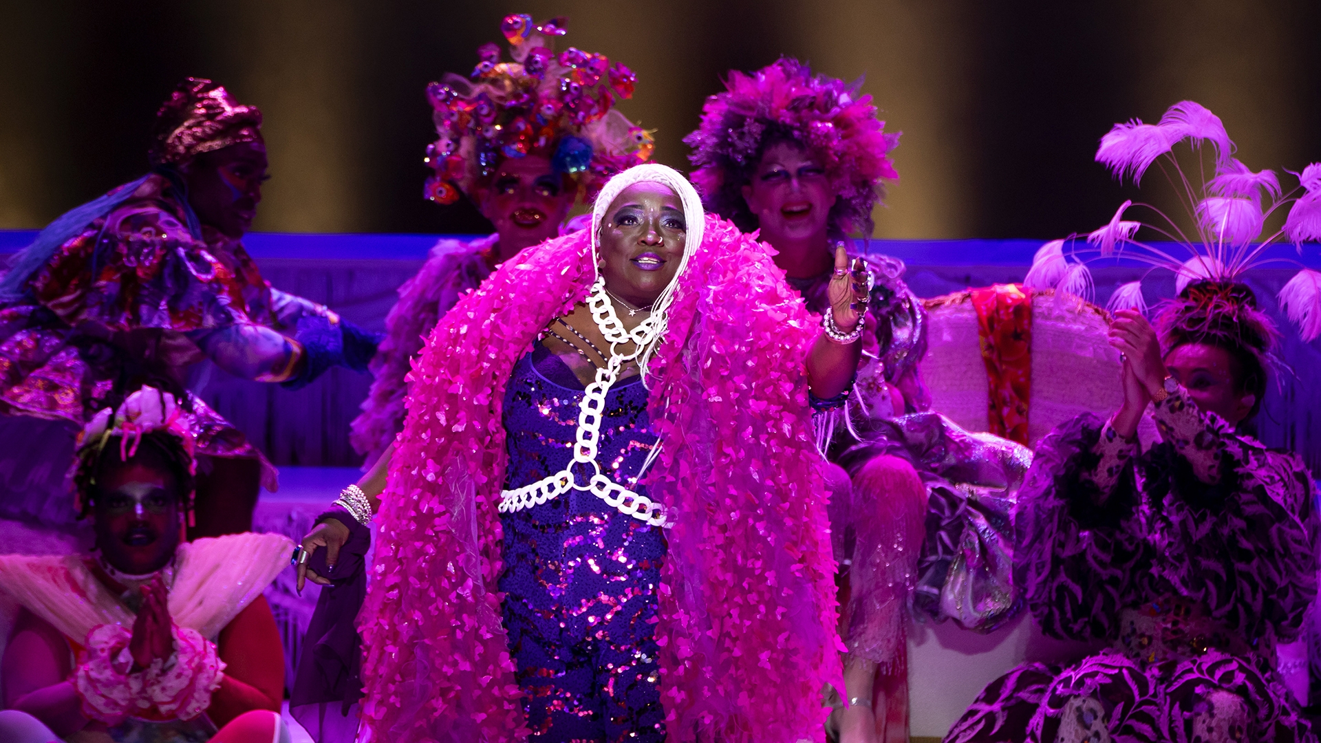 a woman wearing a bright purple sparkling gown and huge magenta boa around her neck performs a song with other performers behind her on stage