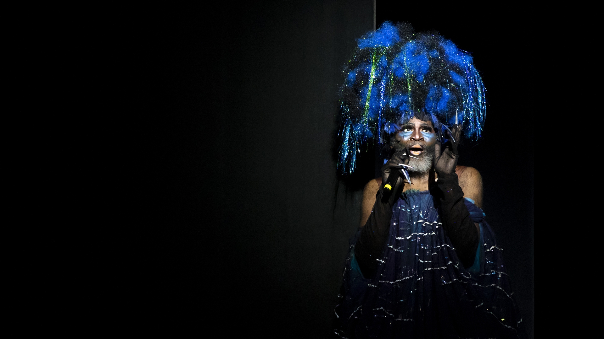 a person with a beard, wearing long black gloves and a giant, fluffy blue and black wig with tinsel hanging from it and a sparkly blue sleeveless dress performs a song on stage