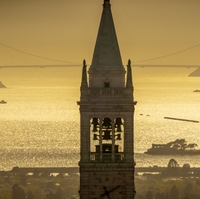 A view of San Francisco Bay from the Berkeley hills. The top of the Campanile is front and center, with shimmering water and the Golden Gate Bridge beyond. The entire photo is cast in a golden light.