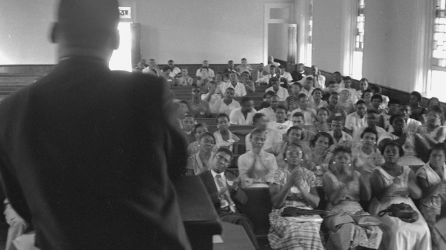 Black and white image of Martin Luther King Jr. with his back to the camera, and standing at a church pulpit speaking to a congregation of churchgoers sitting on church pews.
