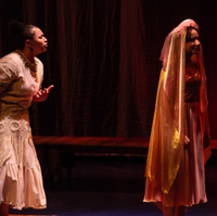 Two actors interact on stage, bathed in a reddish orange light. One actor, wearing a wedding dress and a veil, turns away from the other actor, who is facing her back and appears to be pleading with her.