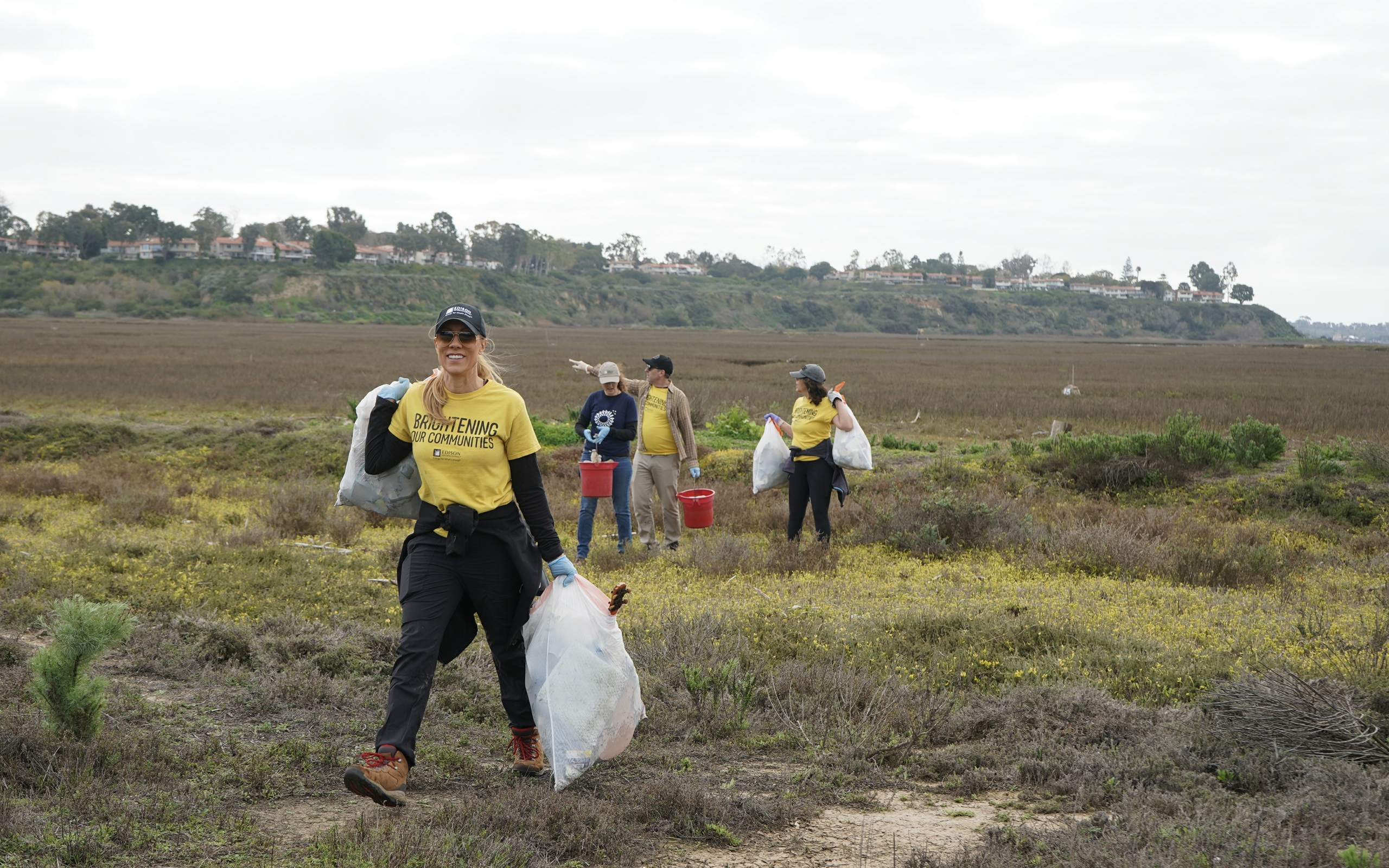Volunteers wearing yellow shirts clean up a grassy field, carrying trash bags and buckets.