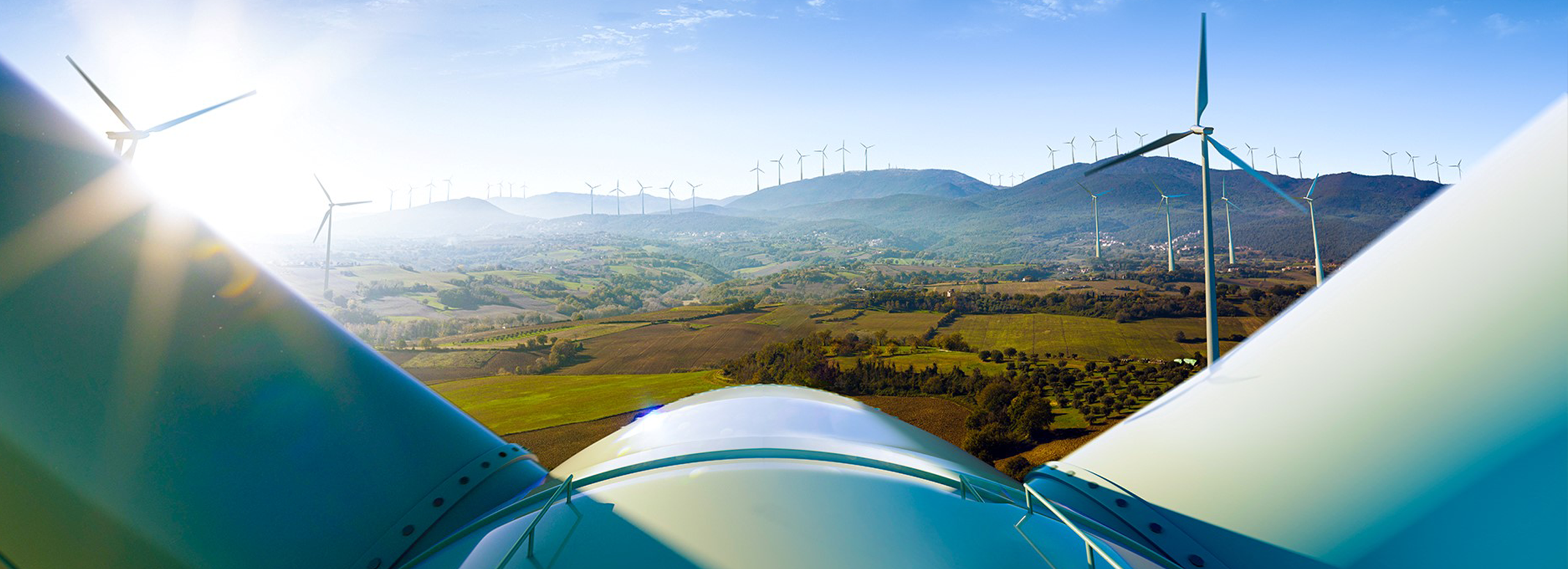Wind turbine blades in the foreground with a landscape of hills and additional wind turbines in the distance under a clear blue sky.