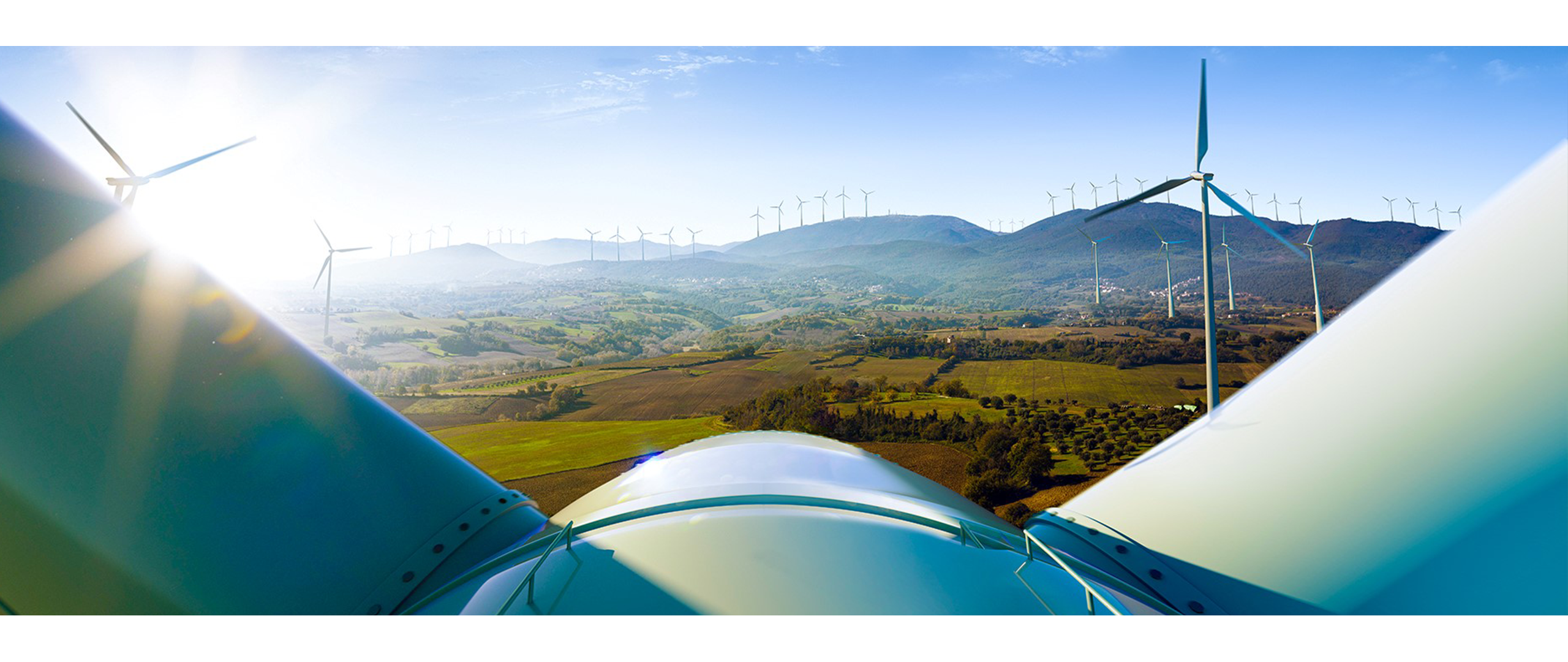 Wind turbine blades in the foreground with a landscape of hills and additional wind turbines in the distance under a clear blue sky.