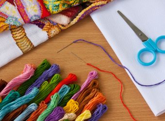 Materials for cross stitch arranged on a desk. Scissors, needle and embroidery thread, and a completed piece.
