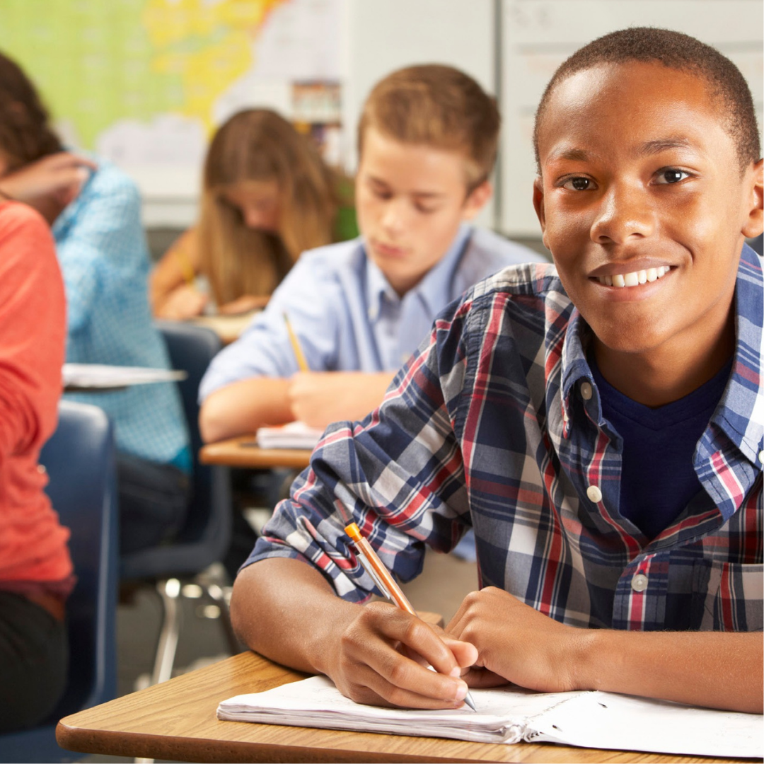 A picture of students in a classroom. An African American boy in a red, white, and blue plaid shirt is sitting at the front of the room in focus. He is looking at the camera and smiling. He is holding a pencil in his right hand and writing on paper. Other students are blurred in the background. 