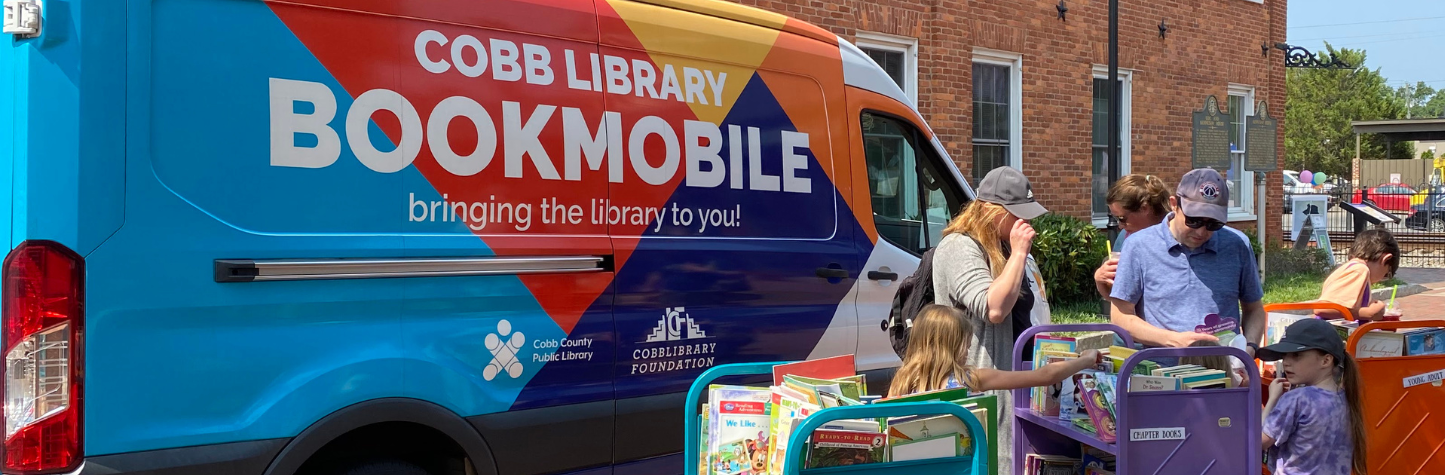 The bookmobile parked in front of a brick building. There are people browsing through books set on colorful book carts in front of the bookmobile.