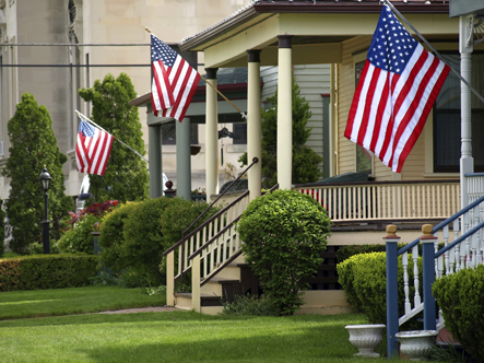 Image features a wide shot of a rural main street residential setting. The homes have American flags hanging in front of them.