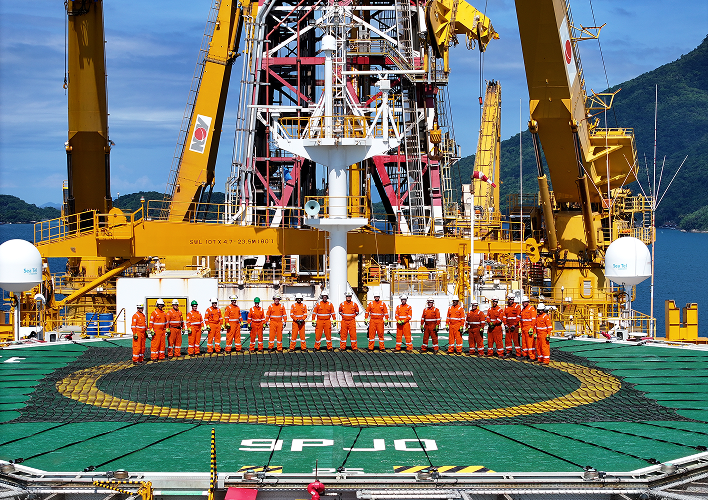 A group of industrial workers wearing orange safety overalls and helmets poses in a straight line on the green helideck (landing pad) of a large drilling vessel or platform. Behind them rises a complex industrial structure with large yellow NOV brand cranes and red and white metal derricks. The sky is blue and there are forested mountains visible in the background.