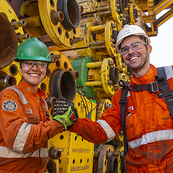 "Dois trabalhadores sorrindo e cumprimentando-se em frente a uma estrutura industrial offshore, com equipamentos de segurança como capacetes e óculos de proteção.