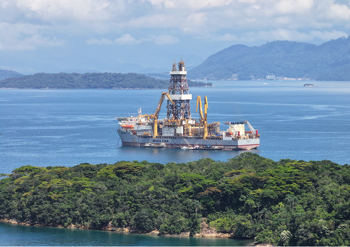Offshore drilling platform near the coast, with prominent structure and visible support vehicles, surrounded by tropical vegetation and open sea.
