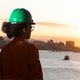 Female worker seen from behind, wearing a green helmet, watching the sunset over the sea with a coastal city in the background.