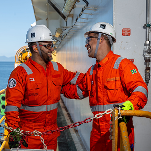 Two workers wearing white helmets and orange overalls talking amicably on an external platform walkway.