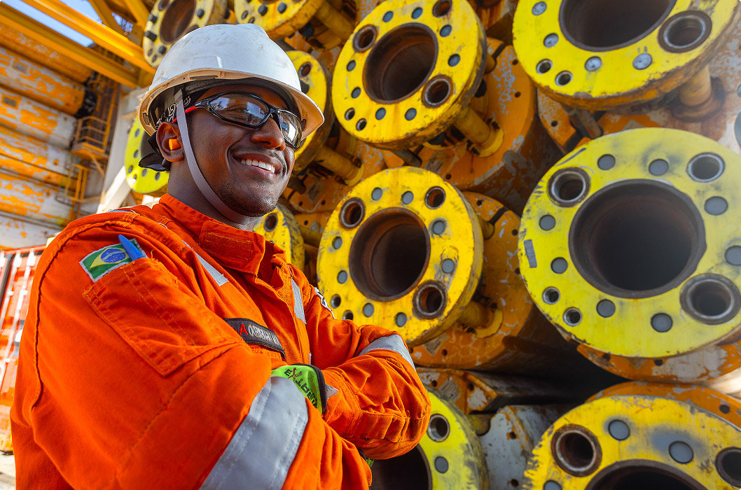 Close-up of a smiling offshore worker, a black man, with arms crossed. He wears a white helmet, safety glasses and orange overalls with the Brazilian flag on the arm. In the background, a pile of large yellow industrial pipes with flange holes.