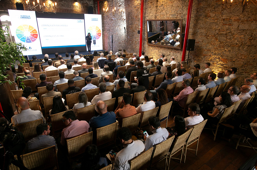 Top view of a corporate event in an auditorium with exposed brick walls. An audience sitting in chairs watches a presentation on an illuminated screen displaying colorful graphics about leadership. A speaker is standing next to the screen.