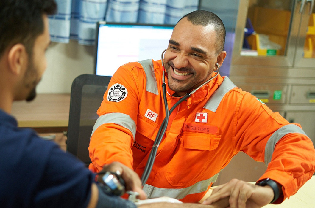 Health professional in an orange uniform, smiling while checking a colleague's blood pressure.