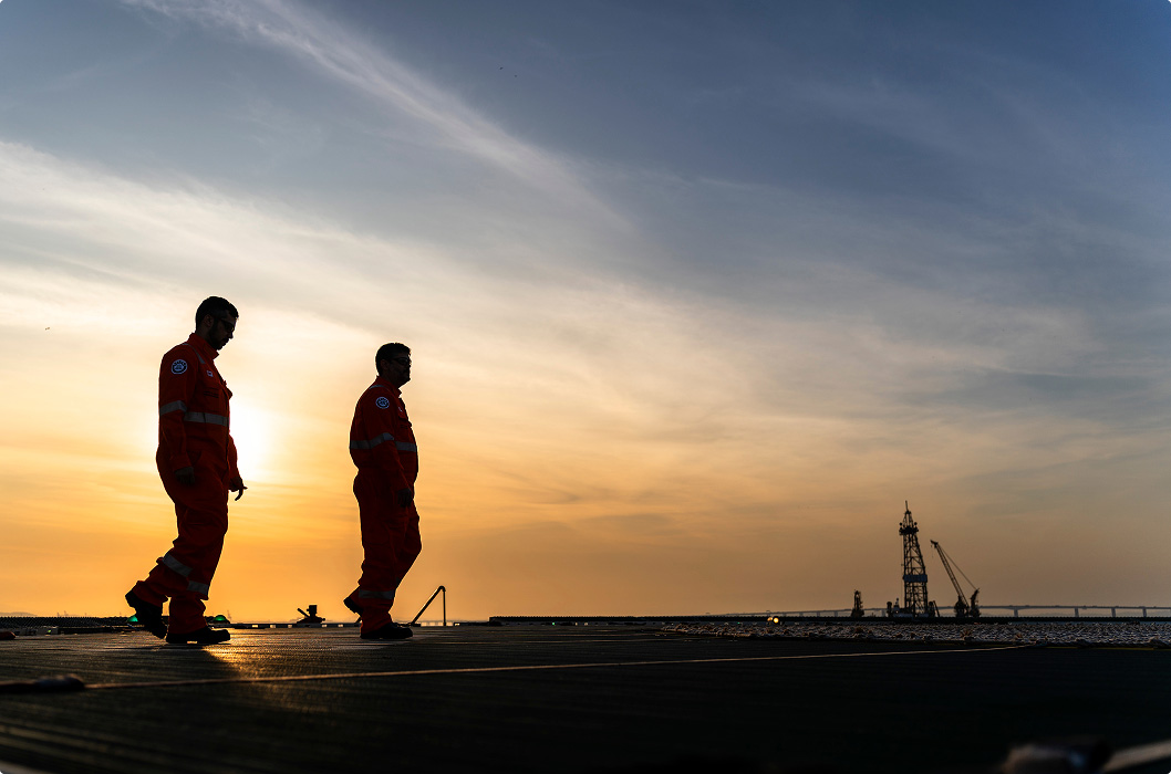 Silhouette of two workers walking side by side on the deck of a platform during sunset. The sky is illuminated in shades of orange and yellow. In the background, on the distant horizon, the outline of a derrick and an extensive bridge over the sea can be seen.