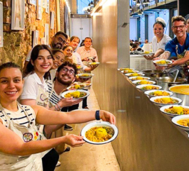 A line of smiling volunteers posing in an industrial kitchen. They wear beige aprons over striped T-shirts and hold plates of food ready to be served. In the background, you can see the Gastromotiva NGO dining hall area.