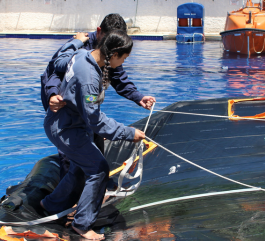 A safety training scene in a pool, where an instructor helps a student walk on the inflatable section of a life raft. The student is carefully guided by the vessel’s safety ropes; both are wearing blue work coveralls