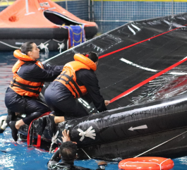 Two women wearing dark coveralls and orange life vests take part in water safety training. They are climbing up the side of a black inflatable raft inside a pool, simulating a rescue drill.