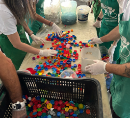 Close-up of volunteers’ hands sorting a large quantity of colorful plastic bottle caps on a table. Participants wear white gloves and green aprons with the campaign logo, organizing the material that will be donated to the One by One NGO.