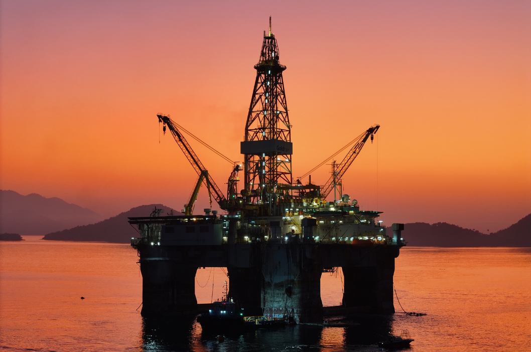 Imposing silhouette of a semi-submersible drilling platform against a sunset sky in vibrant shades of orange and purple. The structure is reflected in the calm surrounding waters, creating a serene and industrial atmosphere.